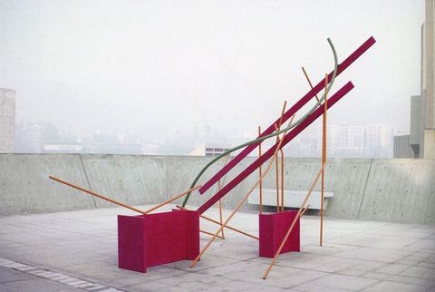 Colour photograph. A metal sculpture made from vibrantly coloured pieces can be seen in an outdoor setting, with the hazy view of a city in the distance.