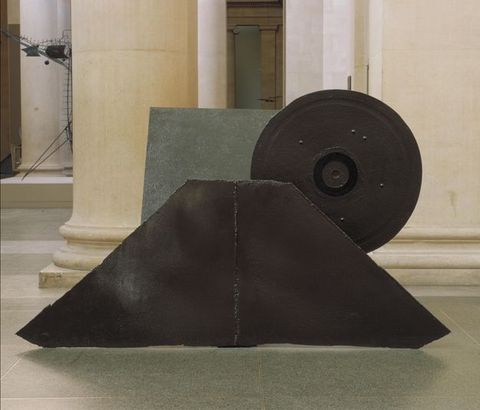 Colour photograph. Documentation of a sculptural metal form made up of bold geometric shapes in an interior space. Several pillars can be seen in the distance.