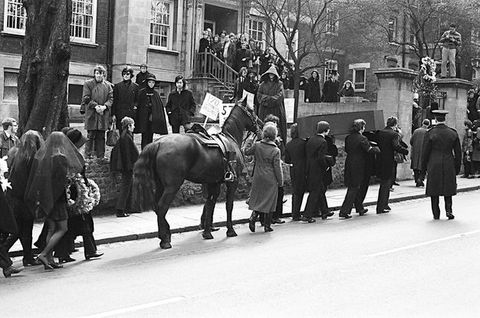 Black and white photograph. A procession of people are seen from behind walking up a road lined by houses, accompanied by a saddled horse. Some members of the group carry circular wreaths.