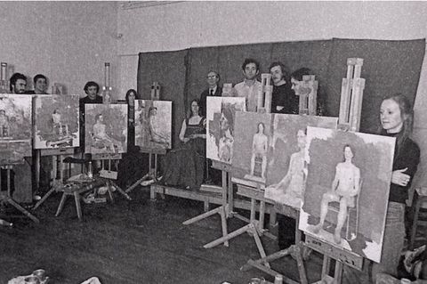 Black and white photograph. Documentation showing an art class, with students standing or sitting behind their easels, each of which is turned out to show similar studies of a nude model.