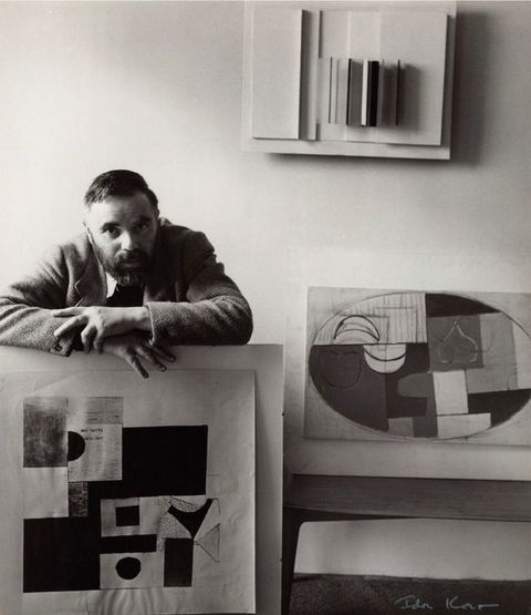 Black and white photograph. A man wearing a smart blazer leans from behind on a board which faces us, bearing an abstract, geometric artwork. Two other similar artworks can be seen around him.