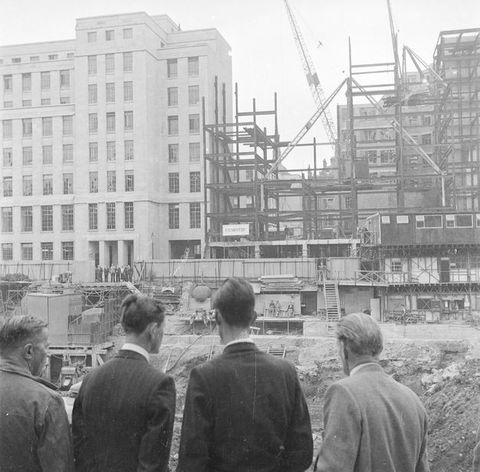 Black and white photograph. A group of four men in smart jackets stand with their backs to us, looking out into a construction site featuring scaffolding and a crane.