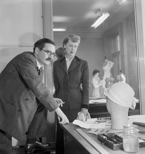 Black and white photograph. A man and woman stand in frontof a table, on which a mannequin' head wearing a hat is seen from behind. A mirror on the wall reveals a woman tailoring clothing on a dummy in the background.