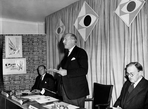 Black and white photograph. Three men wearing smart suits behind a table can be seen from a slight side-on view. One stands in the centre, with mouth parted as if mid-speech, while the other two are seated. Three diamond-shaped posters bearing simple logos are hung on a curtain behind them.