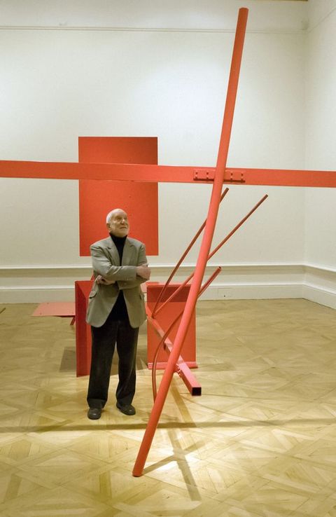 Colour photograph. A man dressed in a pale grey jacket and dark trousers looks up to a partially seen sculpture in front of him, made from bright orange metal. Behnid him is an interior space featuring white walls and pale brown flooring.