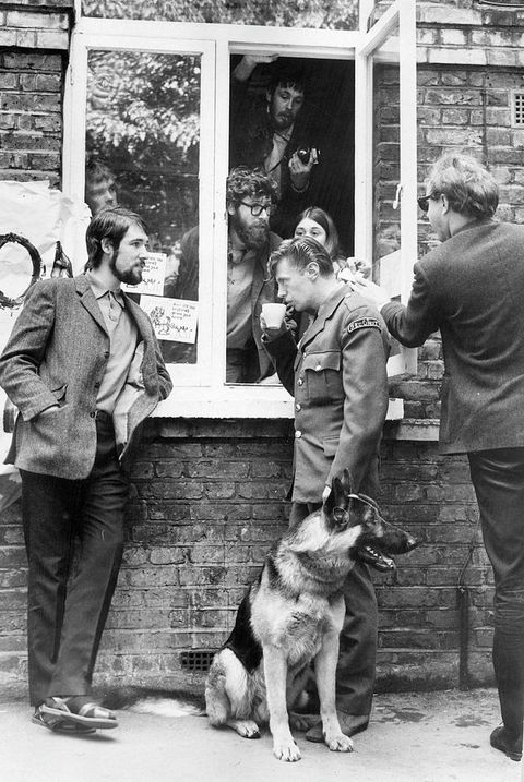 Black and white photograph. Three men in various positions stand on the street in front of an open window. One wears a uniform, sips from a teacup, and is accompanied by a large dog. Other people lean out of the window from the inside. A small section of a hand-painted banner can just be seen to the left, hung on the outside of the brick building.