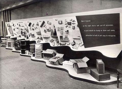 Black and white photograph. An indoor scene showing a wall and floor display to the right of the image. The display features various boxes and barrels arranged along the floor, while the wall shows text accompanied by printed imagery.